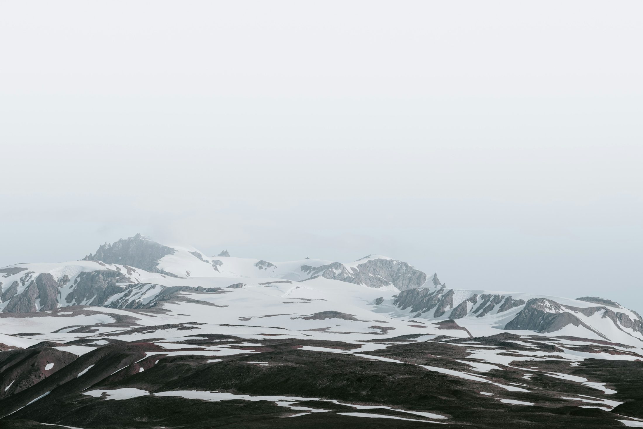 A snow-covered range under flat cloud, bare dark ground in the foreground.